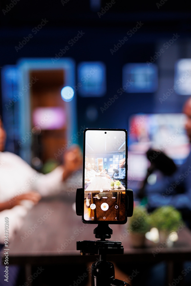 Poster in a neon-lit studio, a vlogger show host is interviewing a guest while a smartphone on a tripod rec