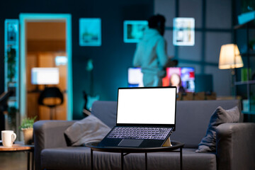 Focus on isolated screen laptop on coffee table in apartment with man in blurry background walking around with headphones on. Close up shot of chroma key device and person in the back doing tasks