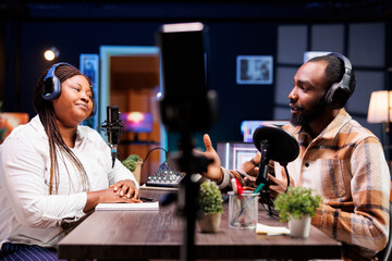 Cohosts filming podcast episode using smartphone on tripod, broadcasting on vlog channel. African american man and woman meeting in studio to record show with mobile phone and talk politics.