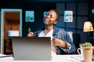 While sitting at her apartment desk, smiling black woman watches comedy film on digital laptop....