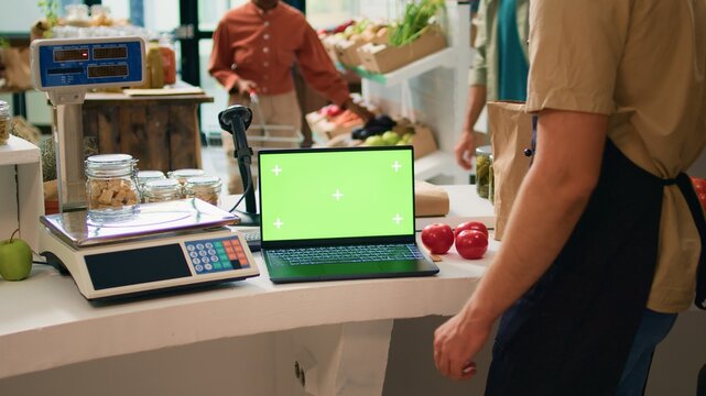 Storekeeper works with greenscreen on laptop, sitting at cash register counter and waiting to serve clients in local zero waste eco store. Seller using blank display with chromakey copyspace.