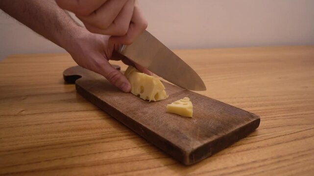 Closeup view of a caucasian man hands slicing a gruyere cheese with a kitchen knife and a wooden cutting board