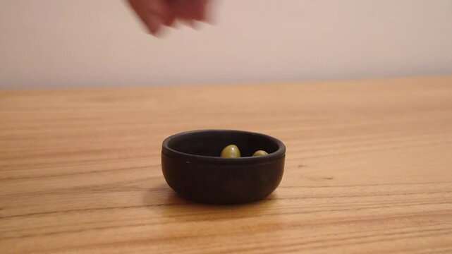 Closeup view of a man caucasian hand placing green olives in a black bowl on the wooden table