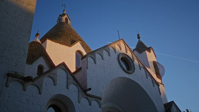 Beautiful church towers in alberobello, puglia illuminated by golden sunlight during twilight, showcasing distinctive conical roofs and whitewashed walls against a deep blue sky.