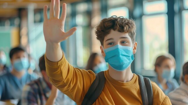 Student in a face mask lifting hand to answer a teacher's question during a health crisis.