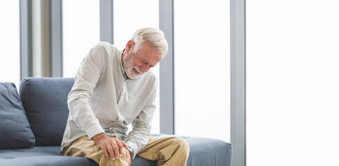 Senior man suffering from knee pain sitting sofa in the living room, Elderly man suffering from...