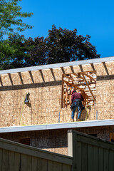 View of new residential home construction over existing wood fence, worker on wall working in window opening of OSB oriented strand board exterior wall, summer construction season
