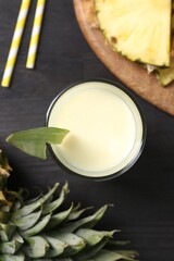 Tasty pineapple smoothie in glass and slices of fruit on black wooden table, flat lay
