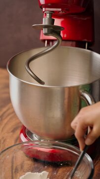 Vertical video, adding all-purpose flour by tablespoons to a stainless steel bowl, red stand mixer