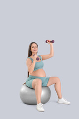 Fototapeta premium Sporty pregnant woman with dumbbells training on fitball against light background