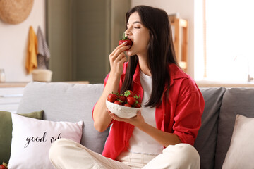 Beautiful young woman holding bowl with fresh strawberries and sitting on sofa in living room