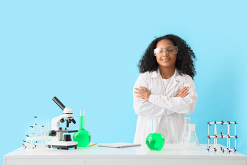 Little African-American schoolgirl with chemical flasks at table on blue background