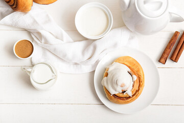 Plate of sweet cinnamon roll with glaze and jug of milk on white wooden table