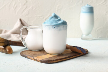 Glass of blue dalgona matcha latte, jug of milk and wooden tray on light table