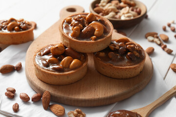 Tasty tarts with different nuts and caramel on wooden board against white wooden background