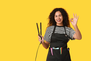 Beautiful young happy African-American hairdresser with hair straightener showing OK gesture on...