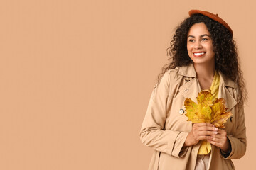 Beautiful young happy African-American woman in stylish outfit with autumn leaves on beige background