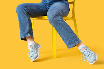 Female legs in stylish sneakers and jeans sitting on chair on yellow background