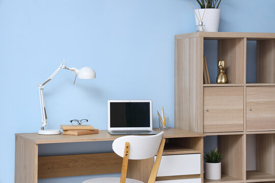 Comfortable workspace with laptop, books and desk lamp near blue wall