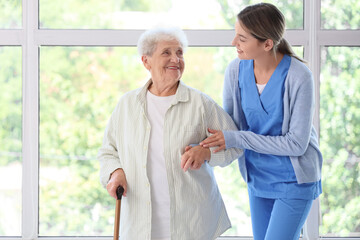 Senior woman with stick and nurse walking near window at home