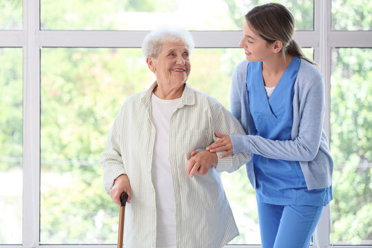 Senior woman with stick and nurse walking near window at home