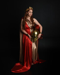 Full length portrait of beautiful female model wearing grecian red silk gold roman robes, historical fantasy goddess costume  holding spartan warrior helmet. Standing pose isolated studio background.