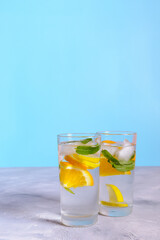 Two glasses of refreshing water with lemon, orange, and mint leaves on a light gray tabletop with a blue background