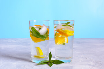 Two glasses of clear, refreshing water with lemon and orange slices and mint leaves, resting on a white surface with a blue background
