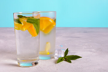 Two glasses of water with orange slices and fresh mint leaves on a gray surface against a blue background