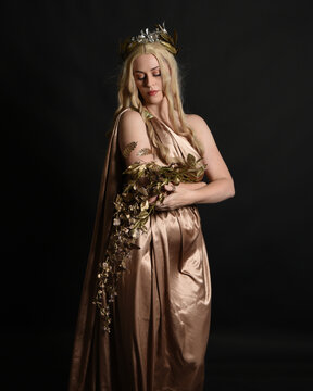 Close up portrait of beautiful blonde female model wearing gold grecian silk toga robes and crown, historical fantasy goddess persephone holding pomegranate fruit. isolated on dark studio background.