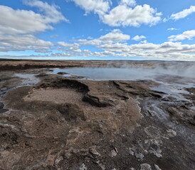 A geothermal landscape in Iceland with steam rising from hot springs against a backdrop of blue skies and clouds. The scene captures the raw and dynamic nature of Iceland's geothermal activity.