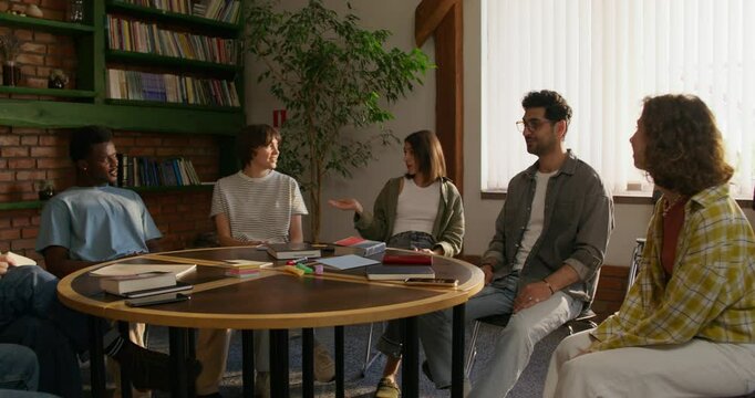 A group of young people are smiling and talking while sitting around a round table indoors. Notebooks and books on the table. Daylight