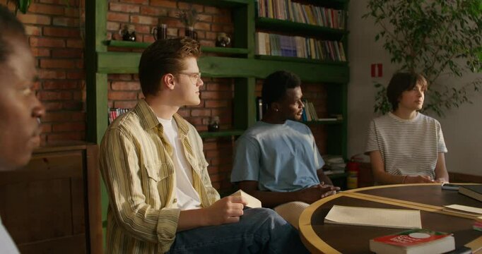 A group of young people are smiling and talking while sitting around a round table indoors. Notebooks and books on the table. Daylight