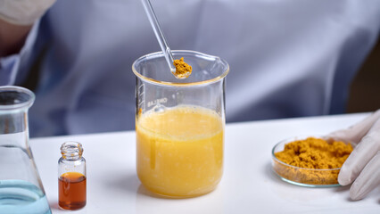 Advertising photo for turmeric cosmetic product in lab scene, was taken from close up front view. The glass spoon of turmeric powder prepare to put into glass beaker, which placed on white table