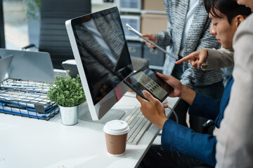 In an office, an Asian team meets at a desk, analyzing financial data on a monitor. They discuss...