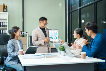 In a boardroom, an Asian team meets at a desk, presenting financial terms and strategies. Executives and employees collaborate, sharing ideas and work guidelines for success during an annual meeting.
