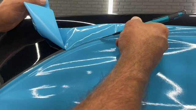 A specialist in the process of work glues the car with an blue vinyl film. Car wrapping specialists cover the car with vinyl sheet or film. Car detailing concept.