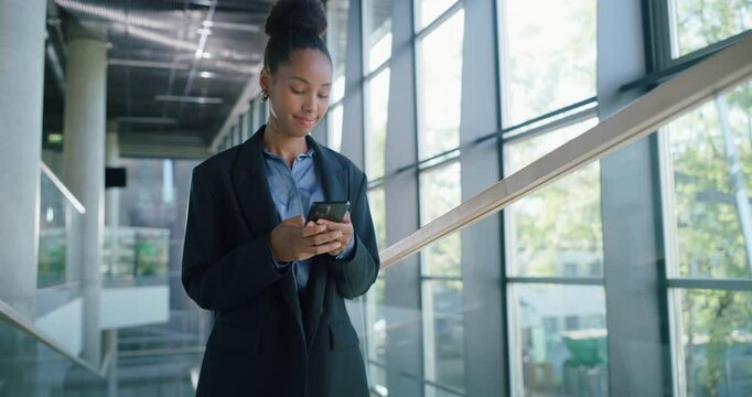 African Female Professional Walking Through a Modern Office Hall, Using Her Smartphone for Browsing Online. Young Woman Connected to the News, Social Network Data and Internet Applications