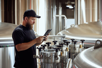 With tablet in hand, brewery technologist checking the brewing machinery, technology control of quality in beer production. Concept of beer, brewery, manufacture, quality control