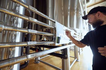 Brewery technologist reviewing brewing process flow, checking labels on stainless steel pipes to ensure accuracy, maintaining quality control in beer production. Concept of beer, brewery, manufacture