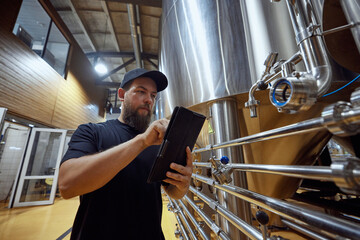 Man, technologist with tablet in hand checking the brewing system, ensuring each step of process meets strict quality standards. Concept of beer, brewery, manufacture, quality control