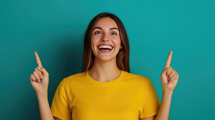 Joyful woman in yellow shirt smiling widely, pointing upwards against a vibrant teal background, exuding excitement and positivity in a cheerful atmosphere.