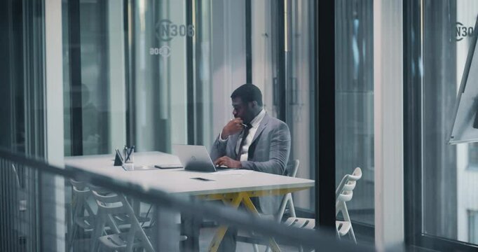 African Manager Focused on Work in a Modern Glass Walled Meeting Room. Company Owner Diligently Using Laptop Computer. Productive Black Male is Dealing with Business Tasks in a Contemporary Workspace