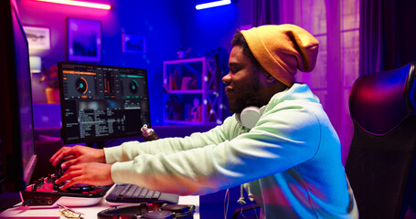 Close up portrait of African male wearing hat with headphones on neck sits in room in front of screen of computer. Handsome bearded face man uses DJ mixer at home.