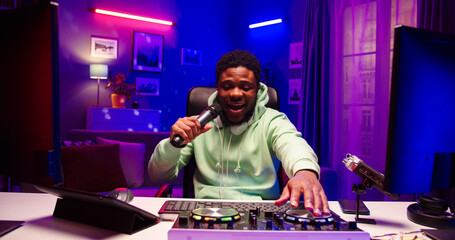 Portrait of happy African male holding microphone in hand sings. Bearded face man turns on karaoke using DJ equipment in LED lighted room.