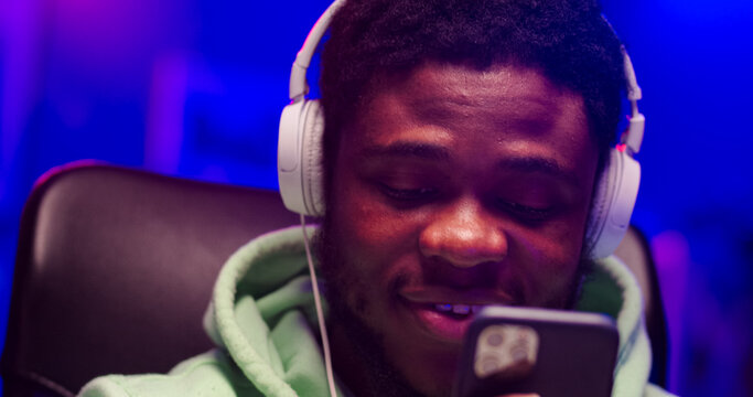 Close up portrait of bearded face African male wearing headphones holds phone in hands. Satisfied man listens to favorite music in room illuminated by LED light.