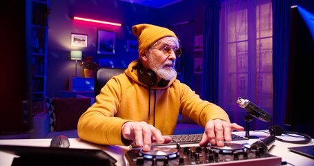 Close up portrait of handsome old Caucasian male wearing glasses with headphones on neck works at creating modern music using DJ mixer. Bearded face man sits in LED lighted room.