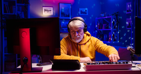 Portrait of handsome old male wearing headphones works using DJ mixer. Bearded face man sits at table with different gadgets in room illuminated by LED light.