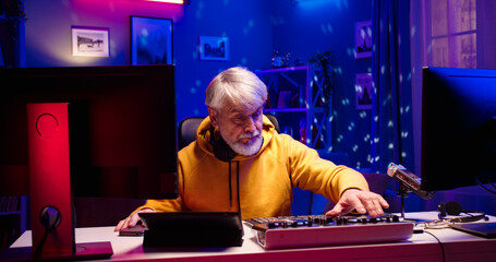 Portrait of handsome old Caucasian male with headphones works at home using DJ equipment. Bearded face man sits at table full of different gadgets in room illuminated by LED light.