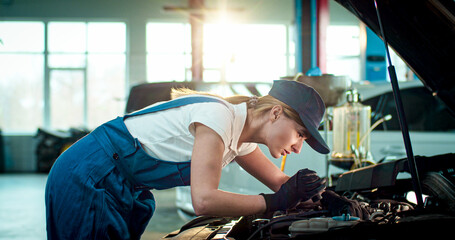 Close up portrait of young Caucasian female wearing working form checks car under hood. Beautiful woman in specialized clothes deftly fixes problems in car.